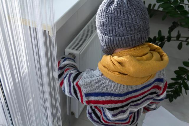 ein Kind mit Schal und Mütze in der Wohnung an der kalten Heizung